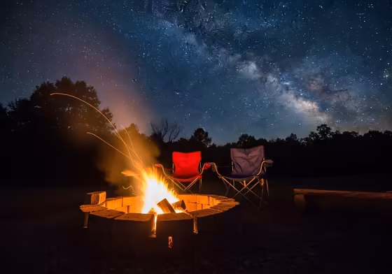 A cozy campfire crackling at a campsite on a clear fall night, with the Milky Way visible in the dark sky above.