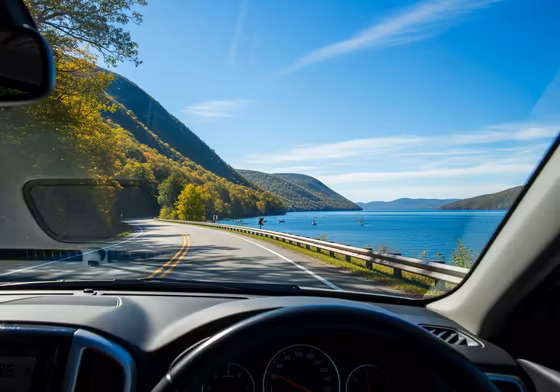 A scenic view of Keuka Lake from the roadside of NY-54A, showing the winding road, the blue water, and the distant hills.