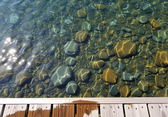 A view looking down from a wooden dock into the exceptionally clear water of Skaneateles Lake, showing smooth, colorful stones on the lakebed.
