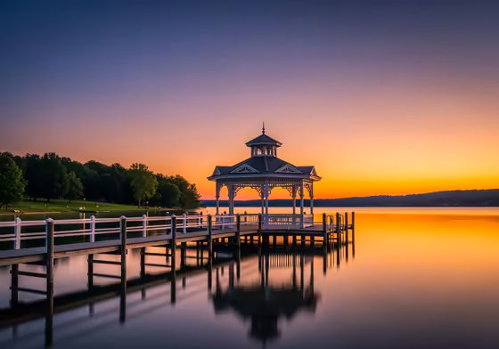 The iconic white pier and gazebo in Clift Park at sunset, with warm golden light reflecting on the calm water of Skaneateles Lake.